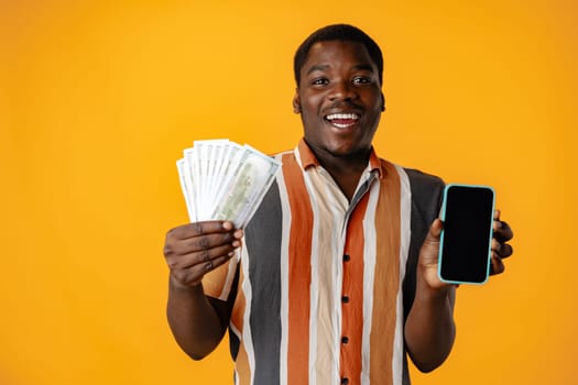 Young african man showing smartphone and dollar banknotes against yellow background