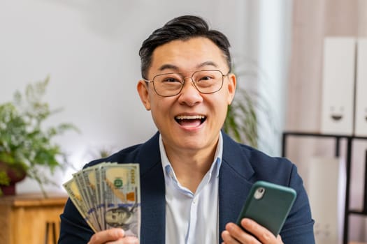 Happy rich Asian businessman counting money cash use smartphone app earning profit success contract