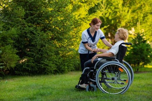 A nurse holds an elderly caucasian woman in a wheelchair by the hand as support. Nurse walks with a patient in the park.