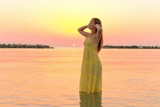 a beautiful girl in a yellow dress stands in the sea straightening her hair against the backdrop of sunrise.