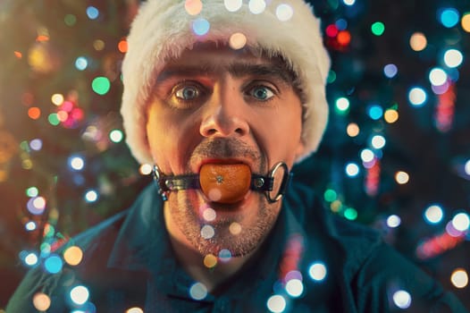 Man with mandarin in his mouth as BDSM sex toys on the Christmas tree background. Concept of fatigue of celebrating New Year's Eve.