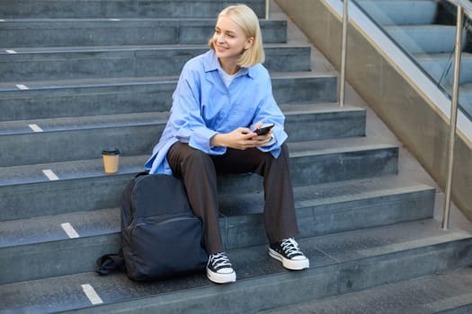 Portrait of woman on stairs, sitting with backpack, using mobile phone, messaging, scrolling social media on lunch break in university.