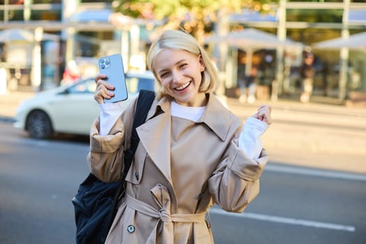 Excited young blond woman with smartphone, standing on street and rejoicing, celebrating victory, triumphing with happy face.