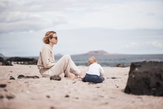 Mother enjoying winter beach vacations playing with his infant baby boy son on wild volcanic sandy beach on Lanzarote island, Canary Islands, Spain. Family travel and vacations concept
