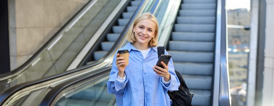 Image of stylish young woman in blue shirt, holding takeaway cup of coffee and mobile phone, standing near escalator and waiting for you, smiling at camera.