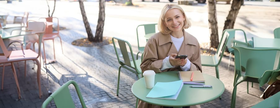 Portrait of cute smiling woman with blond short hair, sitting with documents and mobile phone, looking happy at camera.