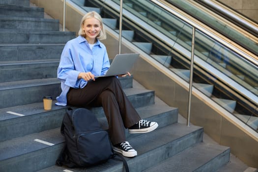 Image of stylish young modern woman, student doing homework, studying outdoors on campus stairs, sitting with laptop and coffee, drinking her cappuccino and connecting to public wifi.