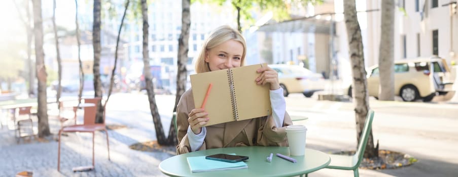 Image of young beautiful woman, student doing homework in an outdoor coffee shop, holding journal or planner, writing in notebook, smiling happily.