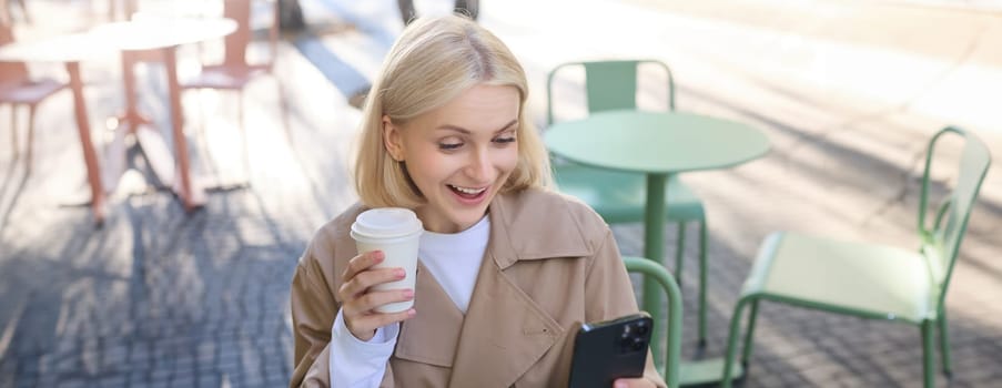 Image of blond female model, looks surprised at her social media account, staring at smartphone with amazed face, drinking coffee outdoors, sitting outside.