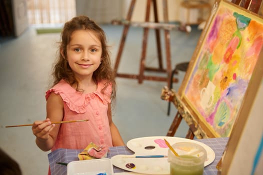 Adorable little child girl artist sitting near picture indoor while learning fine art in creative studio