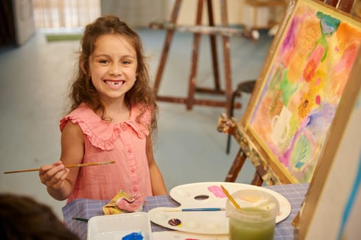 Cute elementary age child girl artist smiles looking at the camera, drawing on canvas while learning fine art in studio