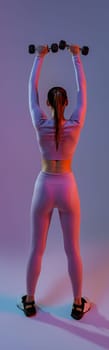 Back view of fitness woman doing exercises with dumbbells on studio background with colored filter