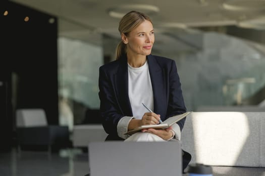Confident female manager making notes in note pad sitting on modern office background