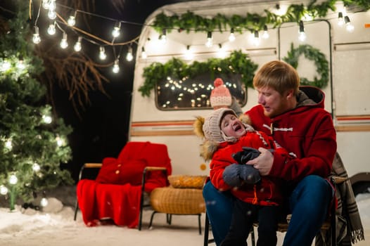 Caucasian red-haired man with a boy in his arms at the mobile home. Father and son celebrate Christmas on a trip.