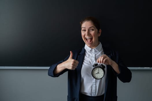 Happy female teacher in pantsuit holding an alarm clock standing at the school board and showing thumbs up.