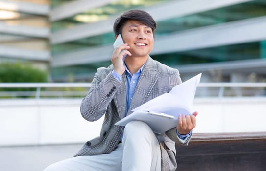 Asian businessman reviews documents and takes calls via smartphone outside