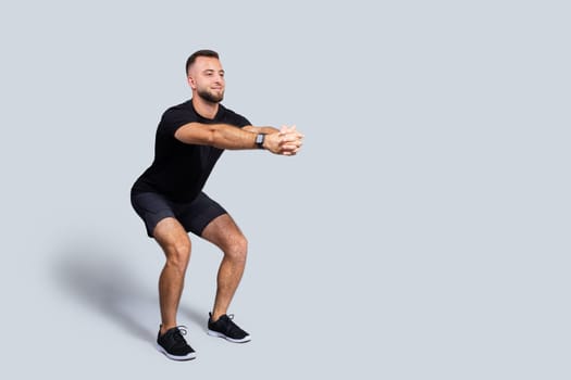Smiling strong millennial caucasian guy with beard in sportswear squat, enjoy sport