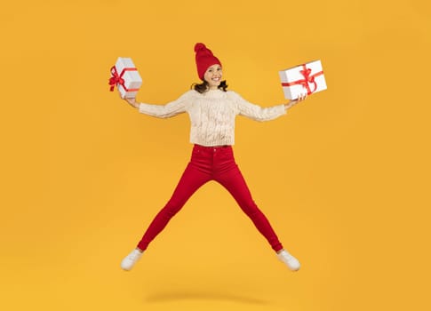 Cheerful woman in knitwear leaps holding two gift boxes, studio