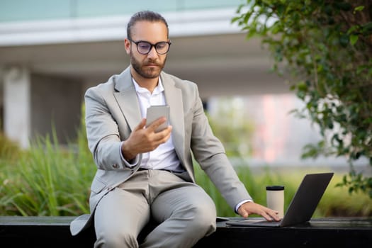 Young professional businessman in suit multitasking with smartphone and laptop outdoors
