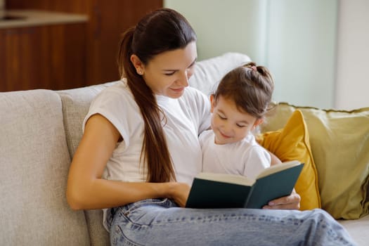 Baby Development. Beautiful Young Mother Reading Book With Her Cute Toddler Daughter While Sitting On Couch At Home, Caring Mom Spending Time With Adorable Female Child, Enjoying Maternity Leave