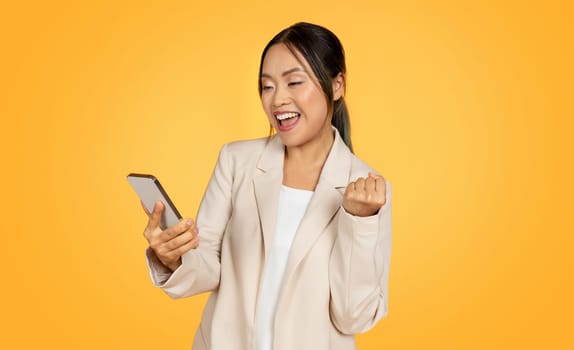 Cheerful young chinese businesswoman in suit with smartphone, enjoy win, celebrate success, rise fist