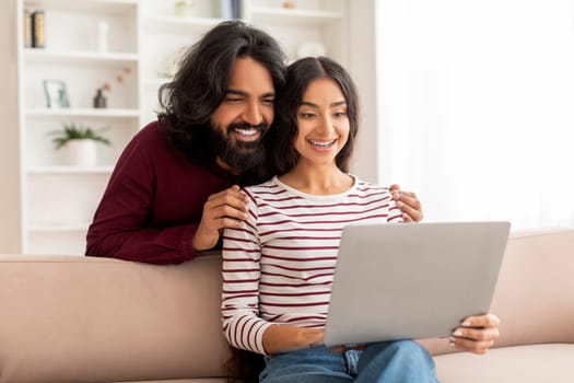 Positive young indian couple have video call with friends, using laptop at home. Happy millennial man and woman sitting on couch, smiling at pc screen, copy space. Telecommunication concept