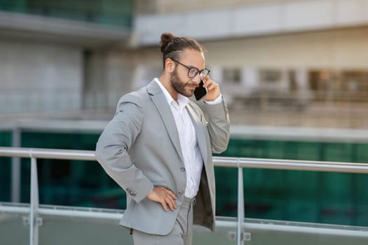 Serious young businessman in suit talking on cellphone while walking outdoors