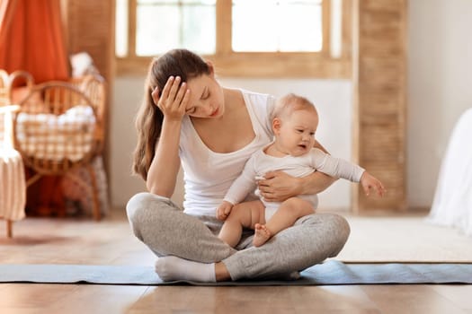 Yoga at home. A young exhausted mother sitting on fitness mat with her crying little baby boy, bedroom interior. Copy space. The concept of fitness with children at home.
