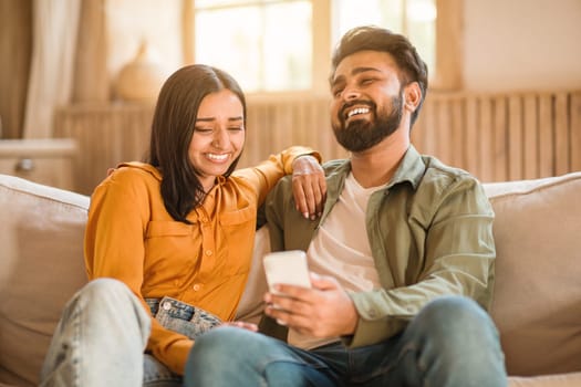 Indian couple laughing and sharing phone on sofa