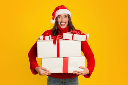 lady in Santa hat holds many Xmas gifts, yellow background