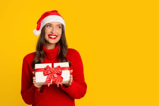 woman in Santa hat presents Xmas gift against yellow backdrop