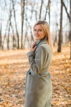 Blonde woman in elegant gray coat walks in the sunny autumn season park. Generation z and gen z youth concept.