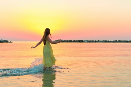 A happy young woman in a yellow dress walking alone along an empty sandy beach at sunset on the seashore.