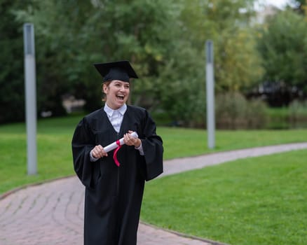 Portrait of happy caucasian woman in graduate gown holding diploma outdoors.