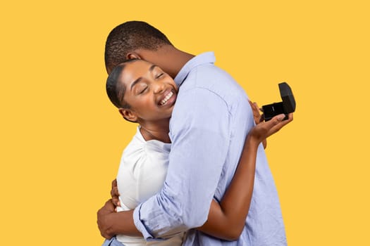 Joyful Black couple hug post-proposal, woman holds ring box, yellow background
