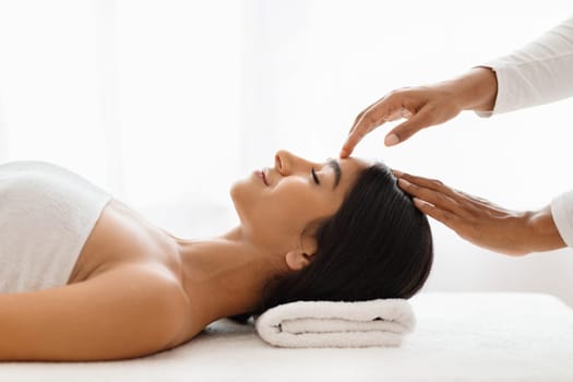 A skilled therapist hands gently kneading the facial areas of a serene, young Indian woman, focusing on her forehead, in an upscale spa setting, side view