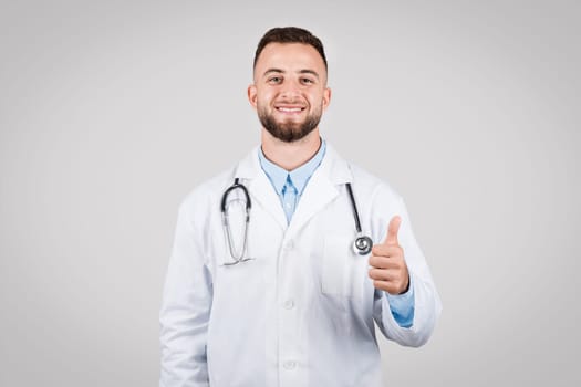 Smiling young doctor with thumb up, white coat, grey background