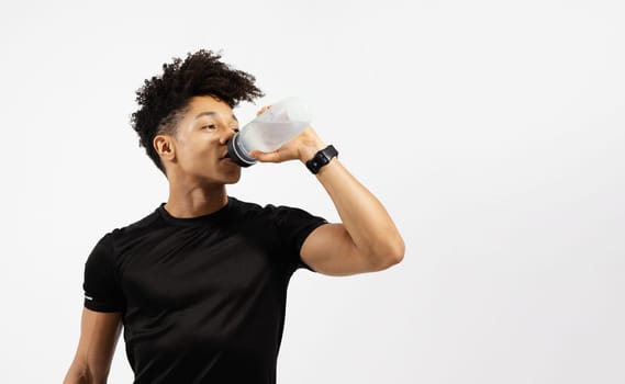 Sporty guy drinking water from fitness bottle posing in black t-shirt, standing over white studio background, near free space for gym advertisement. Fitness lifestyle, hydration