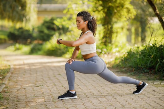 Young fitness woman in park lunging forward, checking heart rate on smartwatch, focused on weight loss and muscle tone, perfect for summer morning workouts and flexibility training outdoors