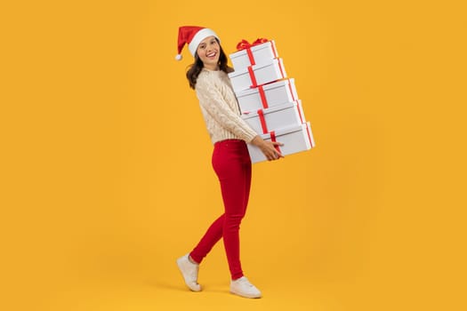 Joyful woman in Santa hat carrying Xmas gifts, yellow backdrop