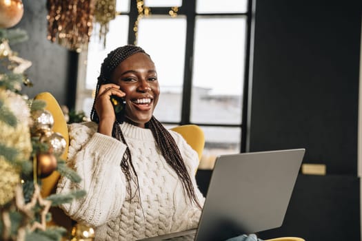 Young African woman using laptop and talking on the phone on Christmas Eve at home