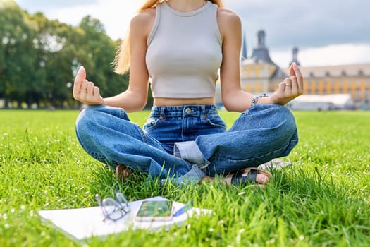 Resting relaxing young teenage girl student meditating sitting in lotus position on grass on lawn, educational building background