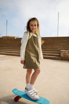 Full length portrait of a Caucasian cute child girl standing on her skateboard on one leg, smiling looking at the camera