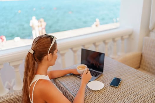 Woman coffee cafe laptop sea. Female freelancer using laptop on coffee break in modern cafe with sea view in the morning