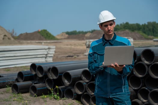 Caucasian male builder in a hard hat stands near the pipes and uses a laptop at a construction site.