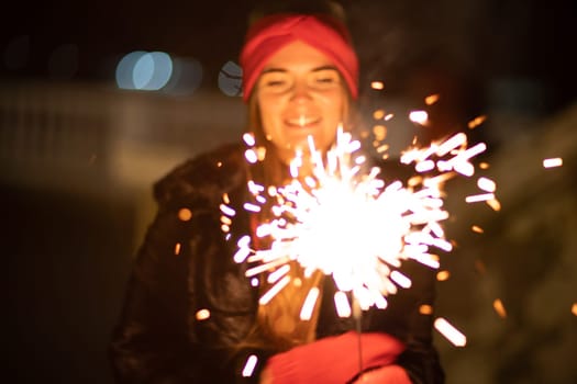Woman holding sparkler night while celebrating Christmas outside. Dressed in a fur coat and a red headband. Blurred christmas decorations in the background. Selective focus