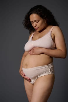 Studio portrait of pretty new mom touching her body with stretch marks and postpartum flaws, isolated on gray background