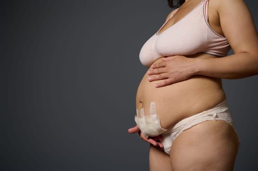 Close-up naked postpartum belly with bandage after cesarean C-section of a body positive woman mother, isolated on gray