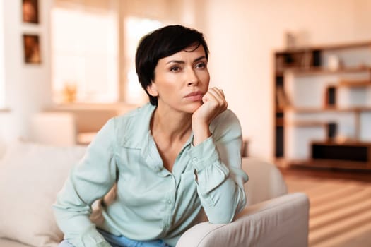 Middle aged woman sitting thoughtfully on her living room sofa