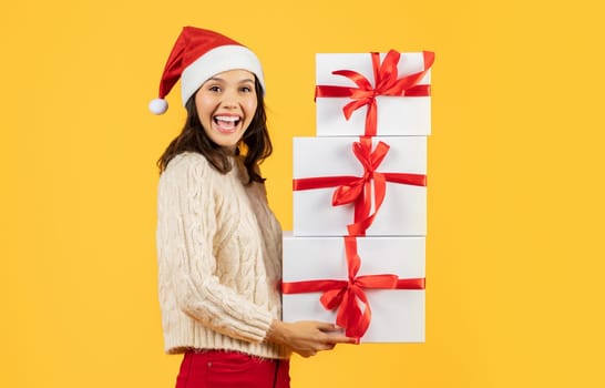 Happy young lady with stack of Christmas gifts, yellow backdrop